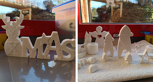 Auf einer Fensterbank stehen zahlreiche, noch reinweiße Gipsfiguren bereit. Links sticht ein großer Schriftzug „XMAS“ hervor, bei dem das X als Rentier mit Geweih gestaltet ist. Rechts daneben drängen sich kleine Wichtel mit Zipfelmützen, Sterne, Tannenbäume und ein Engel mit einem ausgestochenen Herz in der Mitte. Das helle Tageslicht von draußen lässt die glatten Oberflächen der Figuren leuchten.