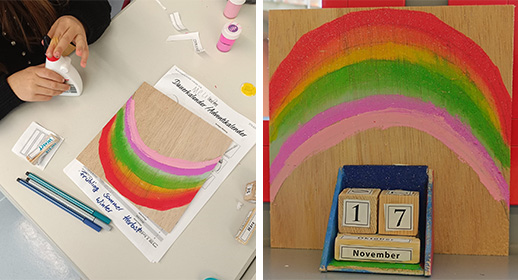 Dieses Bild zeigt die Gestaltung eines Holzkalenders. Links malt ein Kind mit kräftigen Farben einen großen Regenbogen in Rot, Orange, Gelb, Grün und Pink auf eine quadratische Holzplatte. Rechts sieht man das fertige Werk: Vor der Regenbogen-Rückwand steht ein kleines Holzgestell, in dem zwei Würfel mit Zahlen den Tag (die 17) und ein Holzstäbchen den Monat November anzeigen.