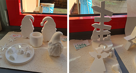 Hier sieht man eine weitere Gruppe von Gipsarbeiten im Detail. Besonders auffällig ist ein weißer Wegweiser, auf dessen Schildern „Welcome“, „Merry“ und „Christmas“ steht. Um ihn herum gruppieren sich verschiedene Wichtel-Figuren mit tief ins Gesicht gezogenen Mützen und knolligen Nasen sowie kleine Gips-Herzen und Sterne, die auf einer schützenden Unterlage verteilt sind.