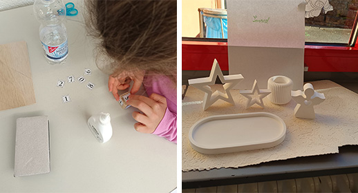 In einer Nahaufnahme sieht man, wie ein Kind konzentriert kleine Papierzahlen auf einen Holzwürfel klebt, um die Datumsanzeige für den Kalender zu basteln. Daneben stehen bereits fertige Gipsformen, darunter ein großer, hohler Stern und ein kleiner Engel. Die Szene vermittelt den Eindruck von ruhiger, präziser Handarbeit an einem hellen Basteltisch.