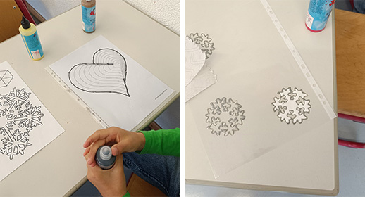 Hier bereitet ein Kind die Dekoration vor. Man sieht die Hände, die eine Flasche mit dunkler Konturenfarbe öffnen. Vor ihm liegen auf dem Tisch Vorlagen für ein plastisch wirkendes Herz und filigrane Schneeflocken unter Klarsichtfolien. Diese dienen wahrscheinlich als Malvorlagen, um die Motive später mit Fensterfarbe oder einer ähnlichen Technik auf die Bastelarbeiten zu übertragen.