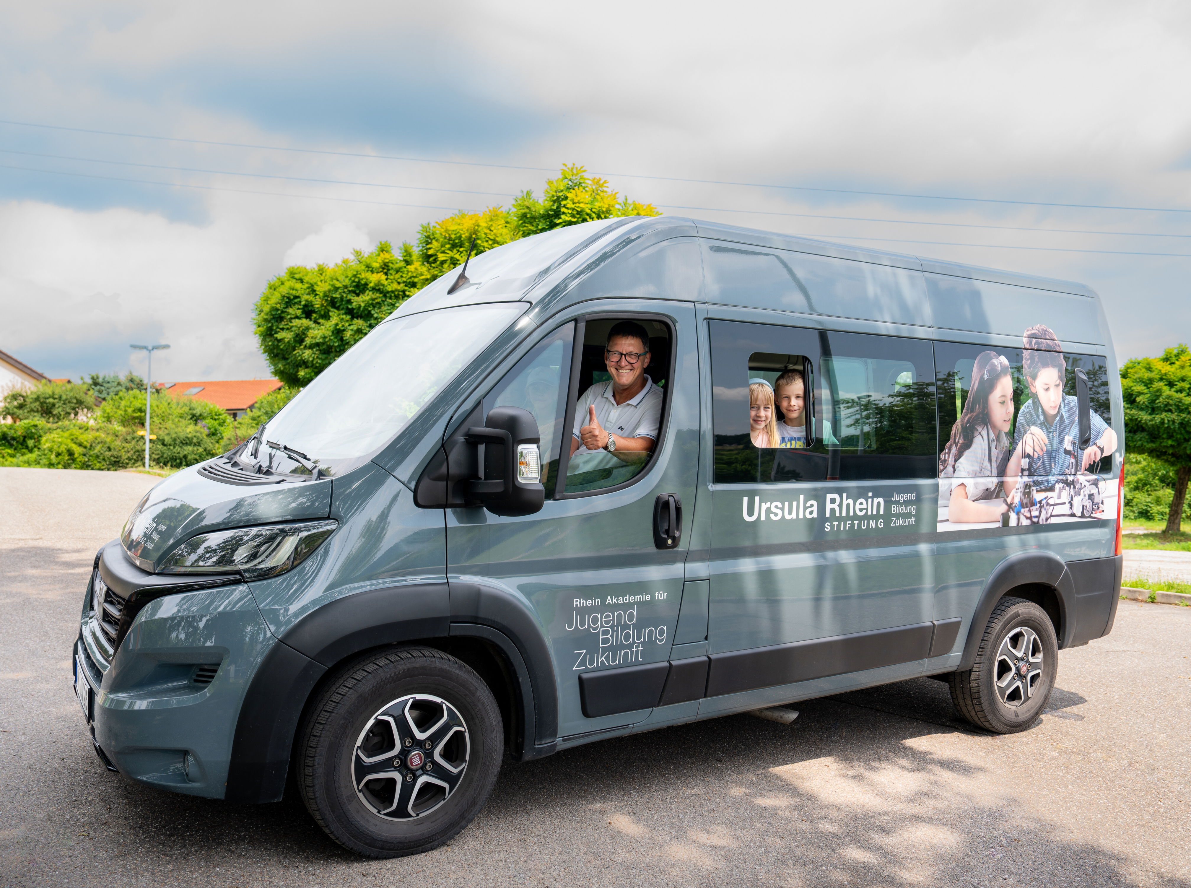 Ein grauer Transporter, der auf der Seite mit „Ursula Rhein Stiftung“ und „Jugend Bildung Zukunft“ beschriftet ist, steht auf einem Parkplatz im Freien. Im Fahrerfenster ist ein lächelnder Mann mit Brille und einem grauen Hemd zu sehen, der den Daumen nach oben hält. Auf der Seite des Transporters sind großflächige Aufkleber mit Bildern von Kindern, die lernen und in der Schule sind, angebracht. Im Hintergrund sind grüne Bäume und ein bewölkter Himmel zu sehen.