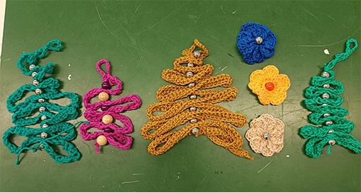 Das Bild zeigt verschiedene Häkelarbeiten auf einer grünen Unterlage. Zu sehen sind vier stilisierte Tannenbäume, die aus bunten Luftmaschenketten in Schlangenlinien geformt und mittig mit aufgereihten Silberperlen fixiert wurden. Sie leuchten in Türkis, Magenta, Goldgelb und Dunkelgrün. Daneben liegen drei kleine Häkelblüten in Blau, Gelb und Creme, die jeweils eine Perle in ihrer Mitte tragen.