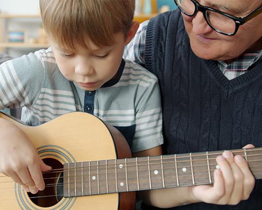 Das Bild zeigt einen kleinen Jungen, der konzentriert auf einer Akustikgitarre spielt, während ein älterer Mann mit Brille und Strickweste (vermutlich sein Großvater) ihm aufmerksam und lächelnd dabei zusieht.