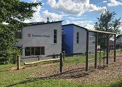 Ein weißes Schulgebäude mit einem blauen Anbau, auf dessen Fassade 'Kämmereischule' zu lesen ist. Im Vordergrund befindet sich ein Spielplatz oder Kletterparcours mit Holzbalken und Spänen auf dem Boden. Grüne Bäume und ein blauer Himmel mit Wolken sind im Hintergrund zu sehen.