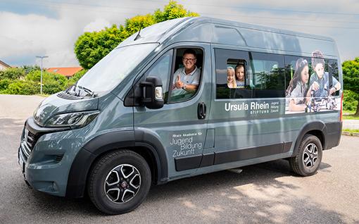 Rhein Akademie Shuttleservice Ein grauer Transporter, der auf der Seite mit „Ursula Rhein Stiftung“ und „Jugend Bildung Zukunft“ beschriftet ist, steht auf einem Parkplatz im Freien. Im Fahrerfenster ist ein lächelnder Mann mit Brille und einem grauen Hemd zu sehen, der den Daumen nach oben hält. Auf der Seite des Transporters sind großflächige Aufkleber mit Bildern von Kindern, die lernen und in der Schule sind, angebracht. Im Hintergrund sind grüne Bäume und ein bewölkter Himmel zu sehen.