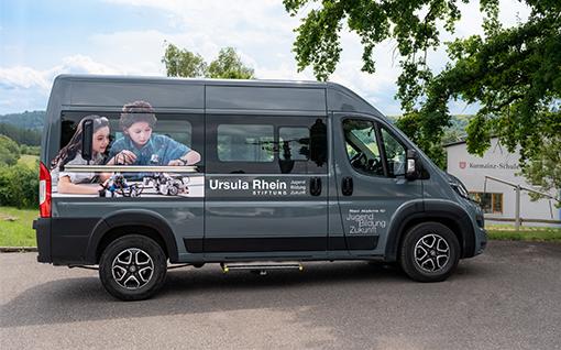 Rhein Akademie Shuttleservice Das Bild zeigt einen grauen Van, auf dessen Seite ein Aufdruck mit zwei Kindern zu sehen ist, die an einem Modell arbeiten. Auf dem Fahrzeug steht der Schriftzug "Ursula Rhein Stiftung Zukunft". Im Hintergrund sind ein grüner Hang und ein Gebäude sichtbar.