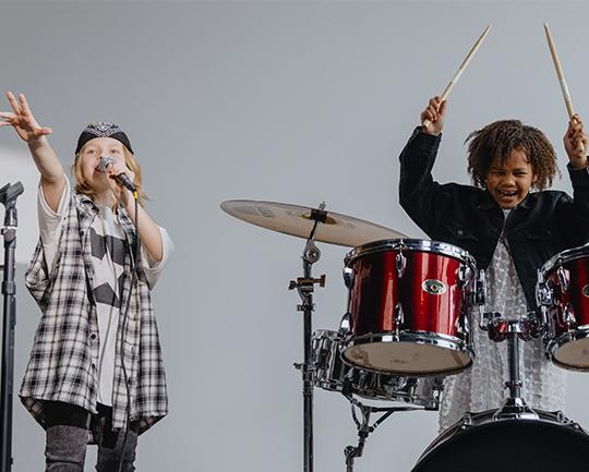 Das Bild zeigt zwei Kinder voller Energie beim Musizieren vor schlichtem grauen Hintergrund: Links singt ein Kind mit Bandana leidenschaftlich in ein Mikrofon, während rechts ein anderes Kind begeistert Schlagzeug spielt und die Sticks in der Luft hält.