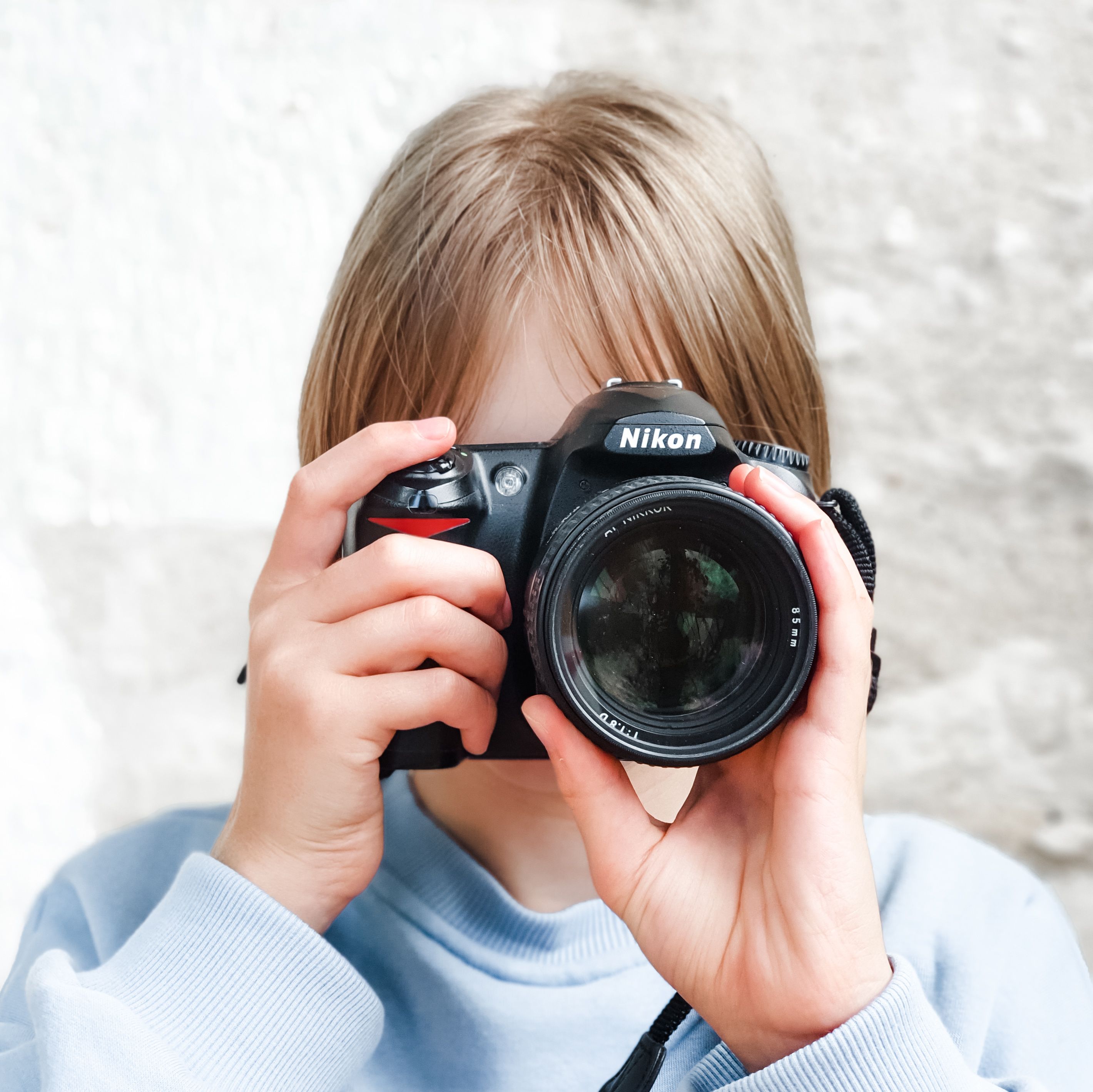 Eine Person mit hellbraunen Haaren und hellblauem Pullover hält eine schwarze Nikon-Spiegelreflexkamera direkt vor ihr Gesicht, um ein Foto zu machen